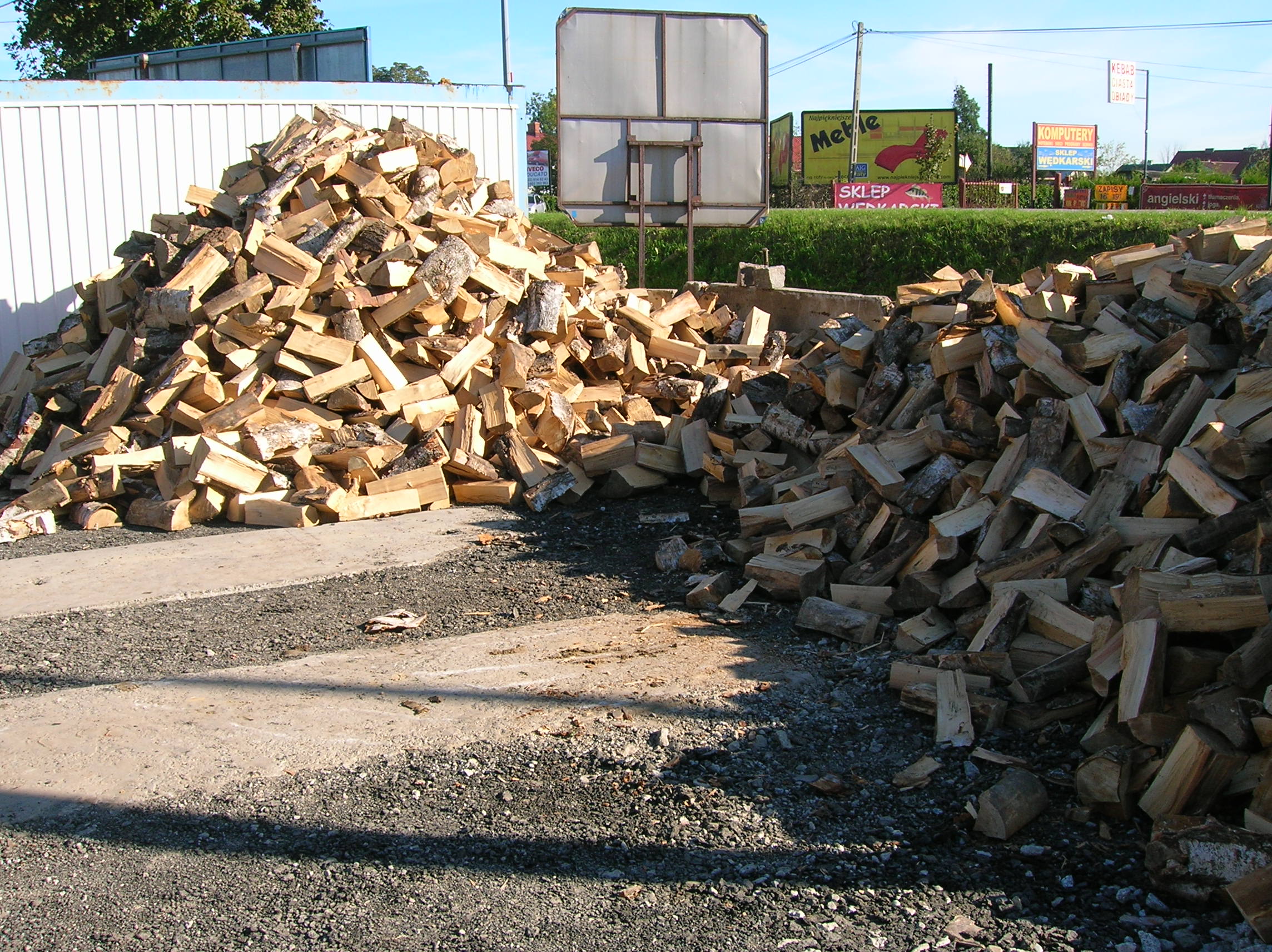 Dwa duże stosy pociętego drewna kominkowego z korą, ułożone na żwirowym podłożu, z widocznymi w tle reklamami i ogrodzeniem.